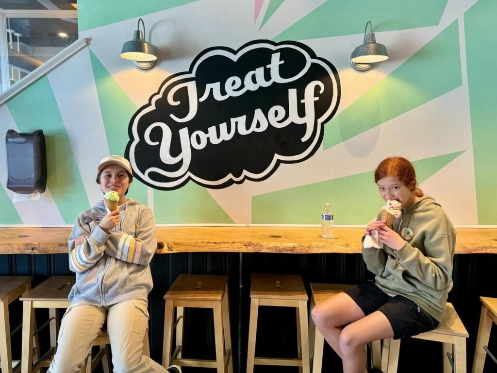 People enjoying ice cream at a cafe with a 'Treat Yourself' wall sign. A colorful geometric background with mint green rays and black lettering.