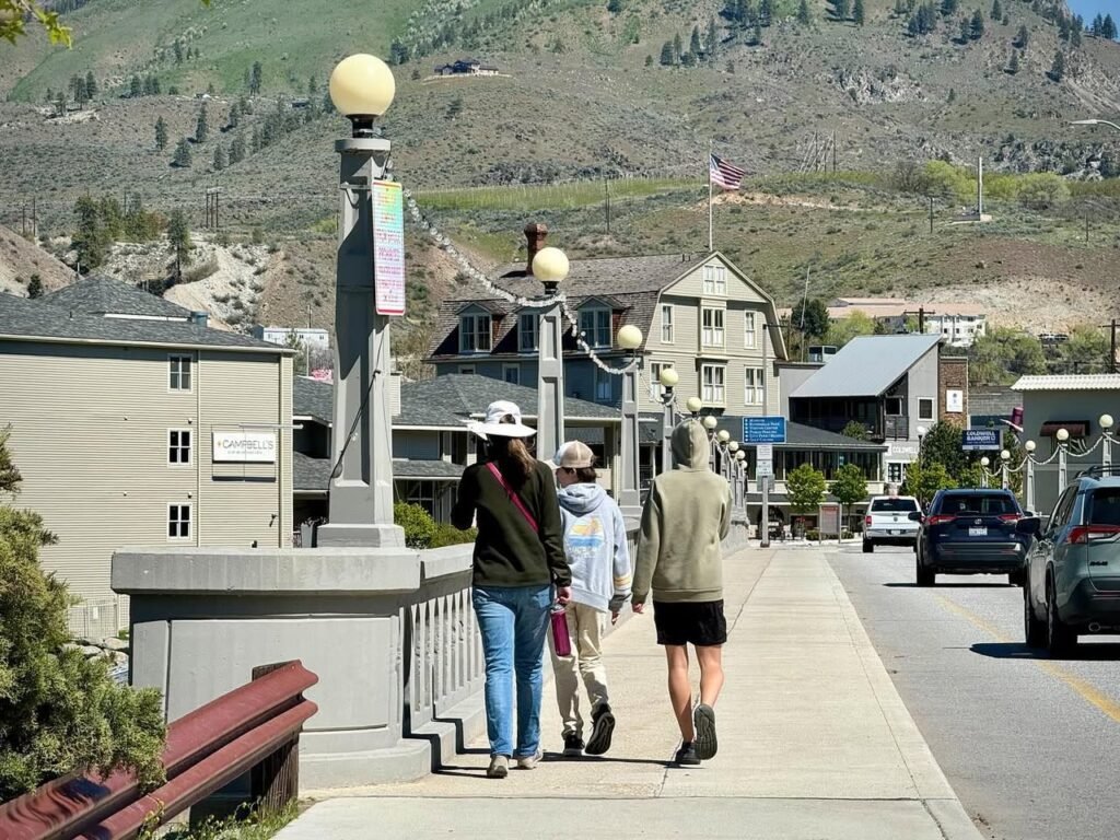 Strolling down a sidewalk in a mountain town. The background features a steep hillside with sparse vegetation and buildings with prominent windows.