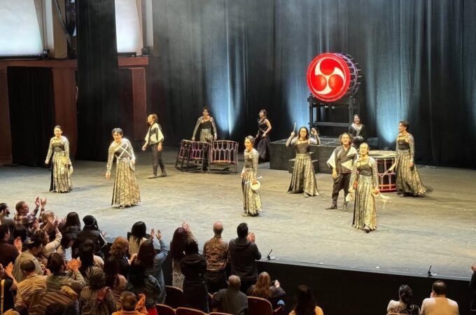 Actors in period costumes perform on a theater stage in front of a live audience, with a prominent red circular prop and dramatic lighting.