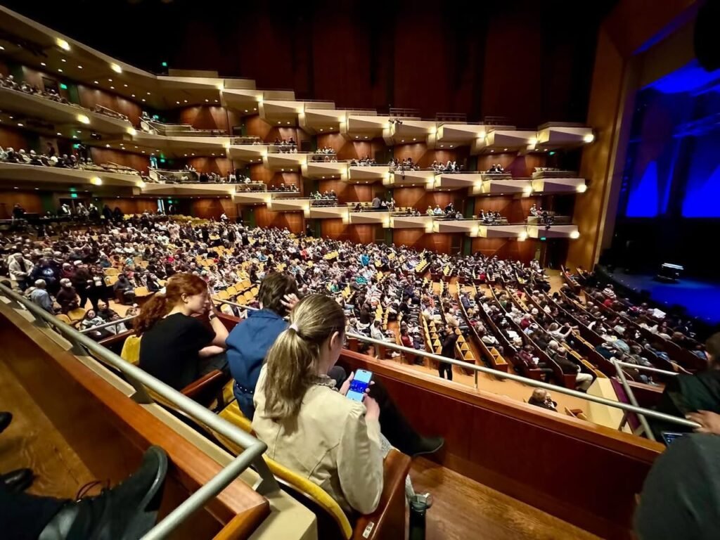 Interior of a live concert venue with tiered seating filled with audience members. The stage is illuminated with blue lighting against warm wooden balconies.
