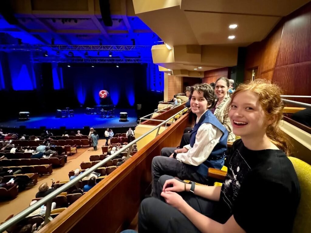 Spectators seated in a theater balcony overlooking a stage with blue lighting.