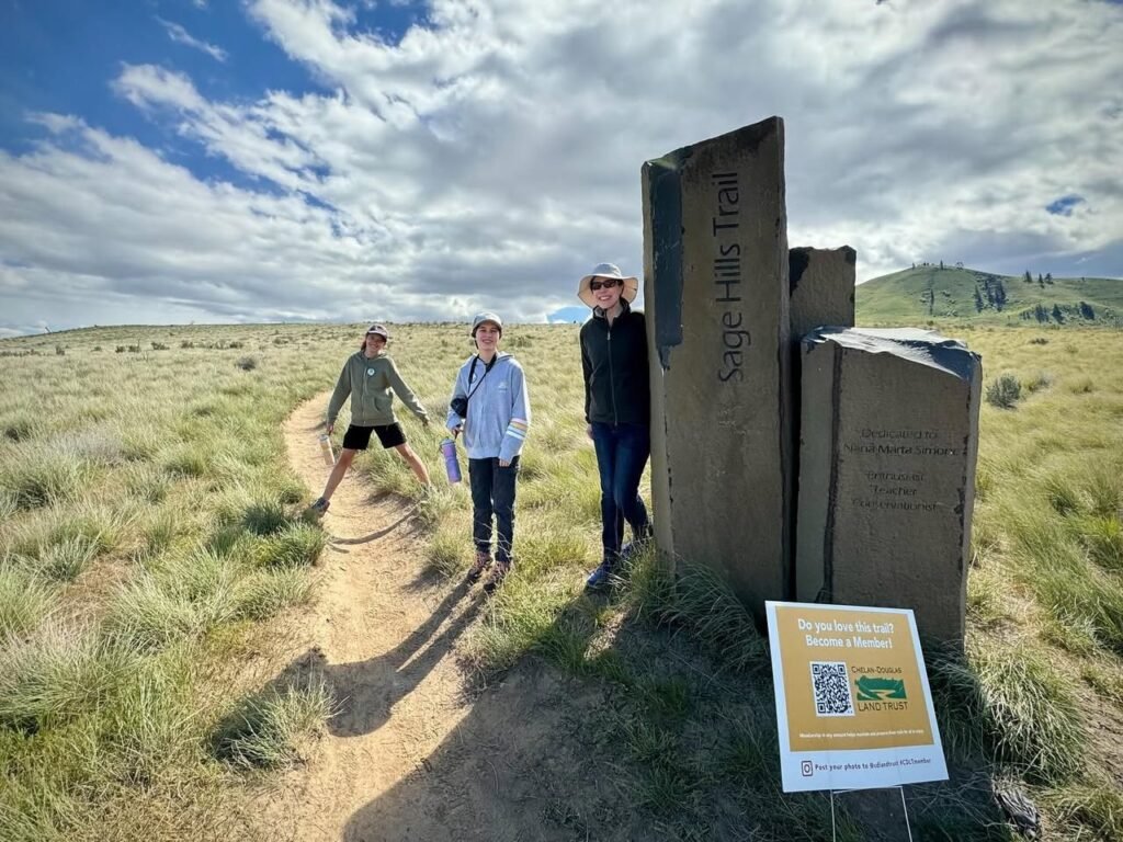 Hikers walk a trail near a stone marker on a sunny day.