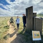 The Aurora Hikers walk a trail near a stone marker on a sunny day.