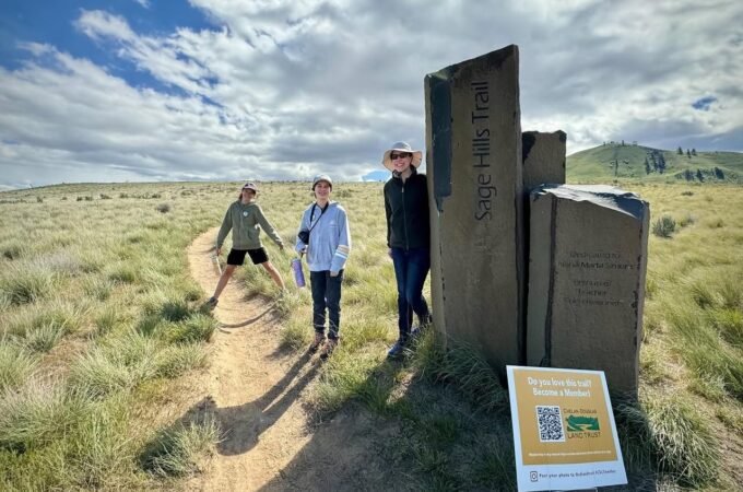 Hikers walk a trail near a stone marker on a sunny day.