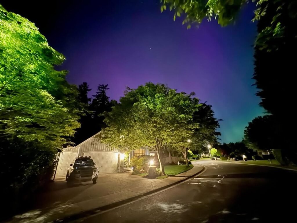 Stunning purple aurora borealis lights up the night sky above a quiet residential street, with illuminated trees and houses reflecting the vibrant glow.