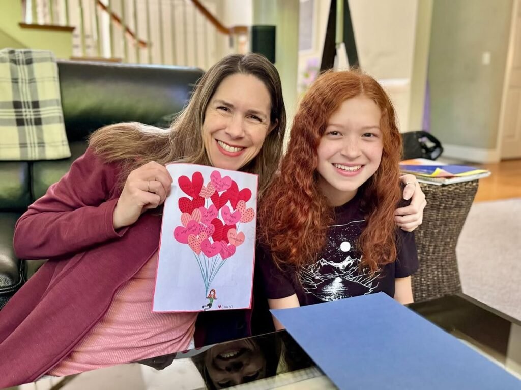 Woman and young girl holding a card with red hearts. A young girl with red hair stands near a blue folder.