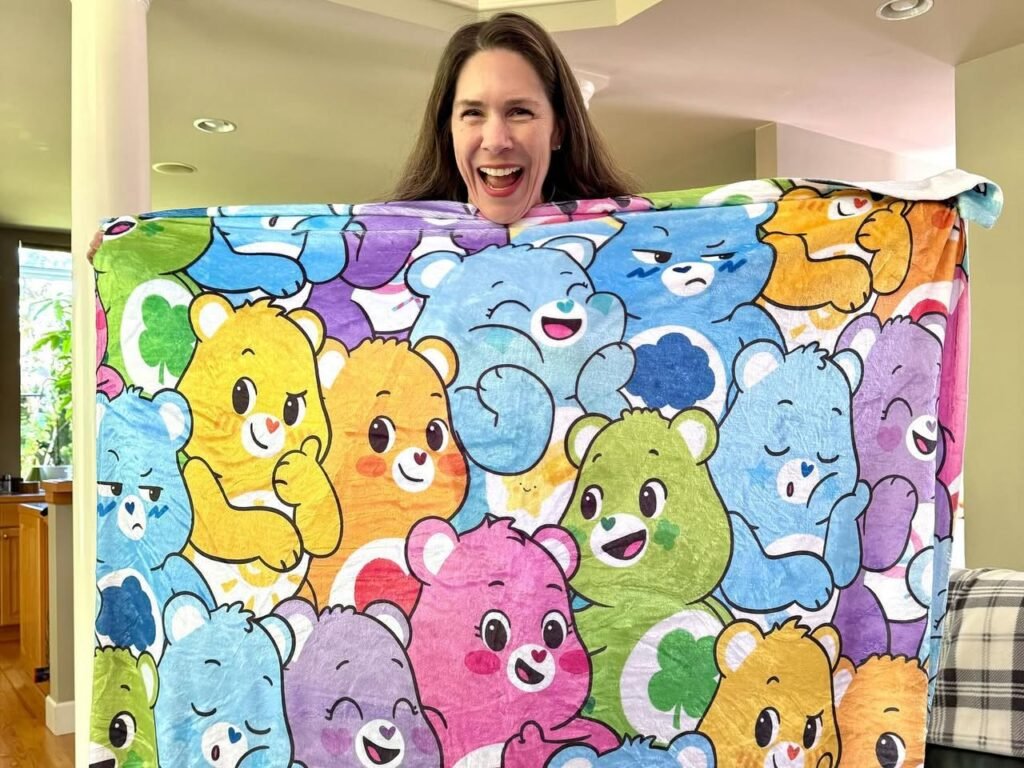 A person smiling while holding up a large red, blue, and green Care Bears fleece blanket in a living room.