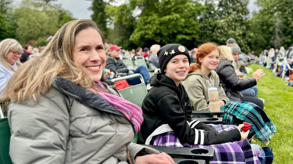A family of four sits together on green stadium seats at an outdoor cultural gathering, smiling warmly against a backdrop of lush trees.