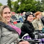 A family of four sits together on green stadium seats at an outdoor cultural gathering, smiling warmly against a backdrop of lush trees.