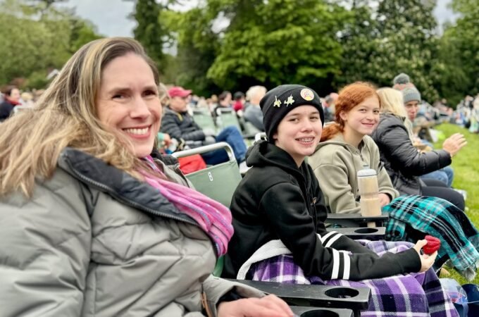 A family of four sits together on green stadium seats at an outdoor cultural gathering, smiling warmly against a backdrop of lush trees.