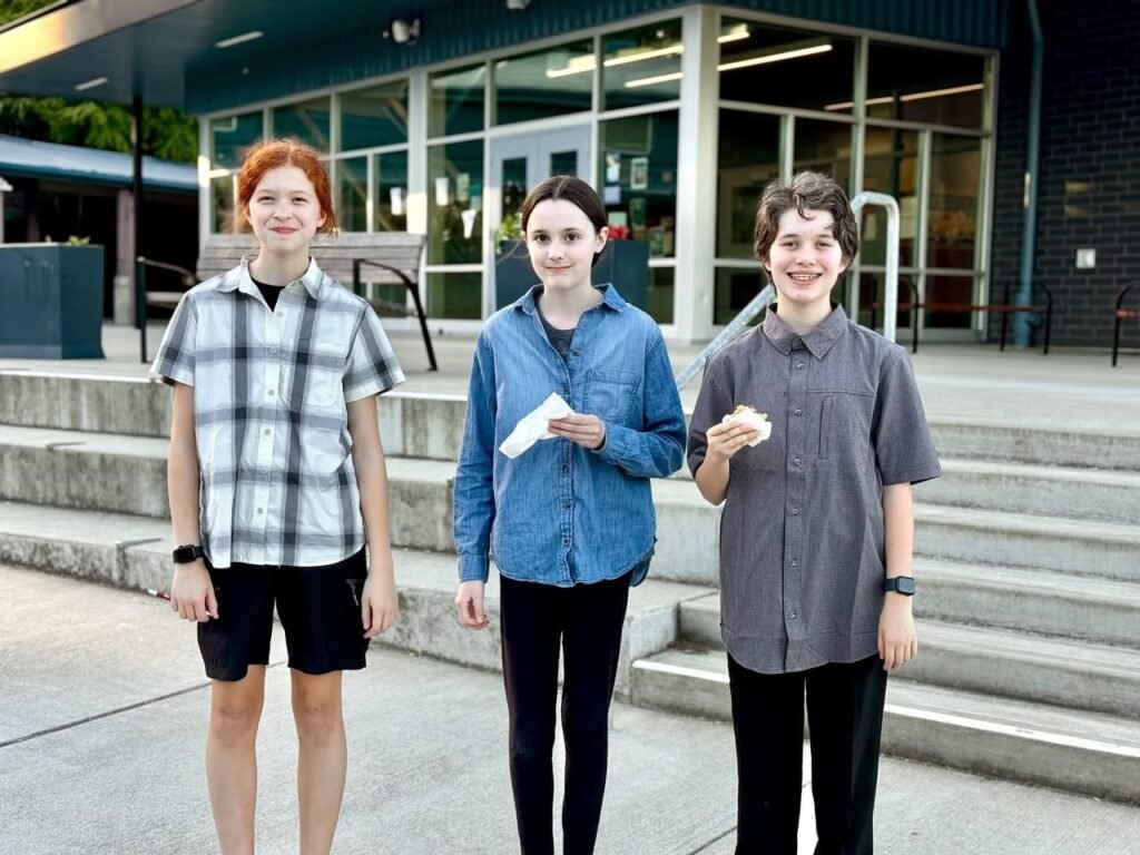 Three individuals standing in casual summer fashion outside a modern building. The photo highlights plaid shirts, denim, and black pants on urban steps.