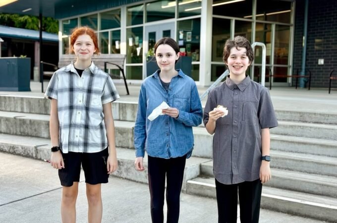 Three individuals standing in casual summer fashion outside a modern building. The photo highlights plaid shirts, denim, and black pants on urban steps.