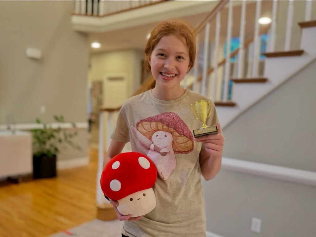 A smiling girl holds a small trophy and a red mushroom plush toy. She wears a t-shirt with a matching mushroom and fairy design in a home hallway.