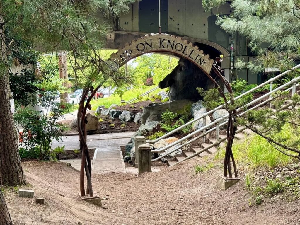 A rustic wooden archway reading "Bear on Knollin" stands at the entrance to a natural trail with stairs and a bear statue nearby.