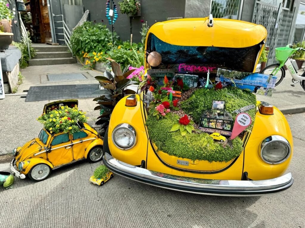 A yellow VW Beetle and a smaller matching planter filled with moss and flowers create a whimsical display on a city sidewalk.