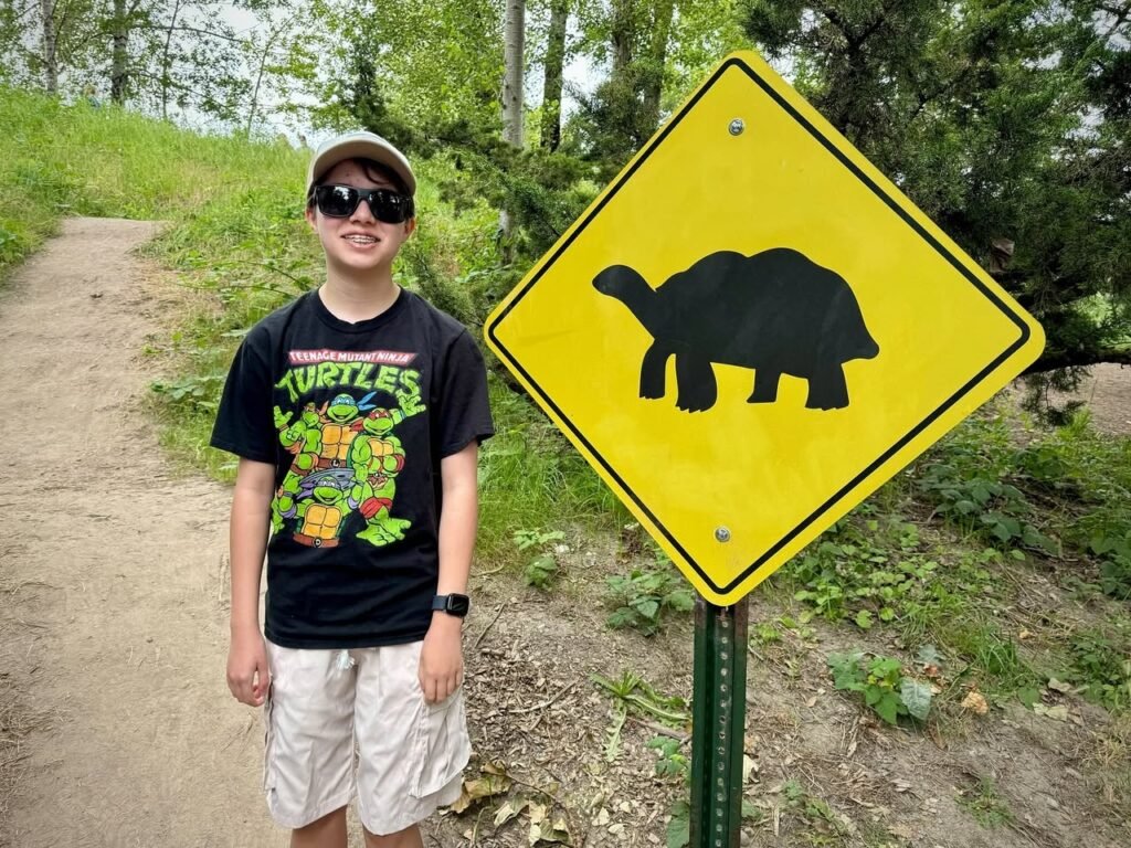 A smiling person wearing sunglasses and a Teenage Mutant Ninja Turtles t-shirt stands next to a yellow diamond road sign featuring a black silhouetted tortoise. The scene is set on a dirt hiking trail surrounded by lush green foliage.