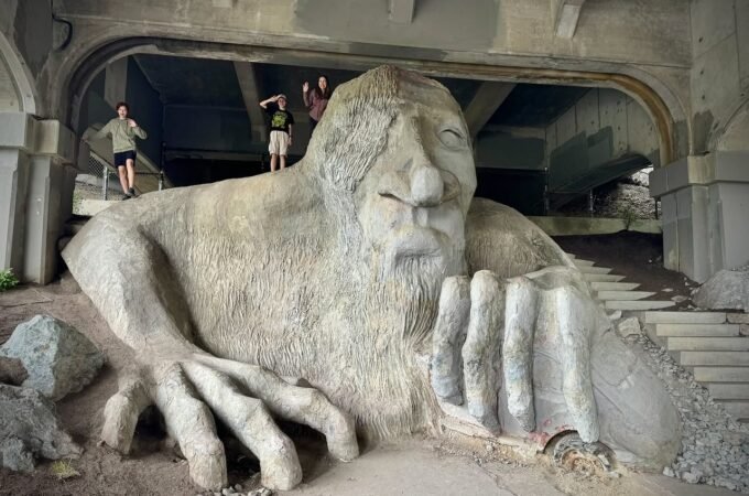 A detailed grand concrete sculpture of a bearded man emerges from the earth beneath stone archways.