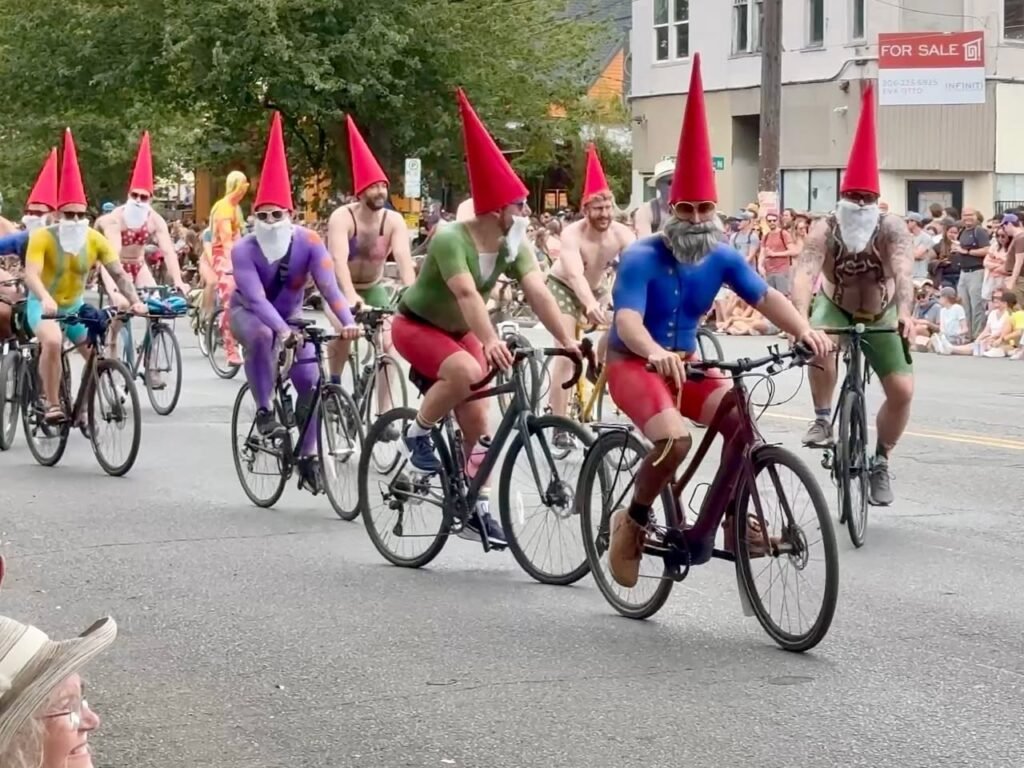 Participants in colorful gnome costumes ride bicycles through a city street with spectators lining the sidewalks.
