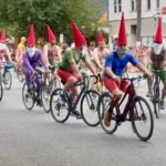 Participants in colorful gnome costumes ride bicycles through a city street with spectators lining the sidewalks.