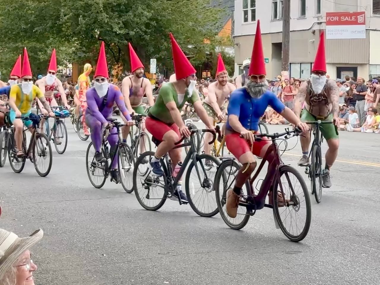 Participants in colorful gnome costumes ride bicycles through a city street with spectators lining the sidewalks.
