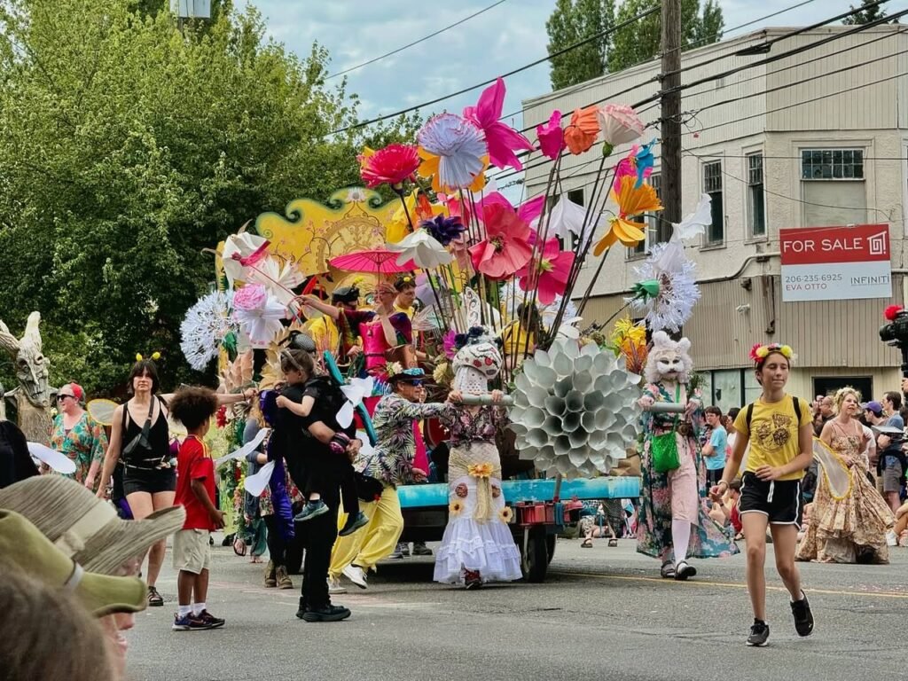 Participants in colorful costumes march behind a float decorated with large paper flowers and lace details during a street parade. Spectators line the roadside under a partly cloudy sky near a building with a red for sale sign.