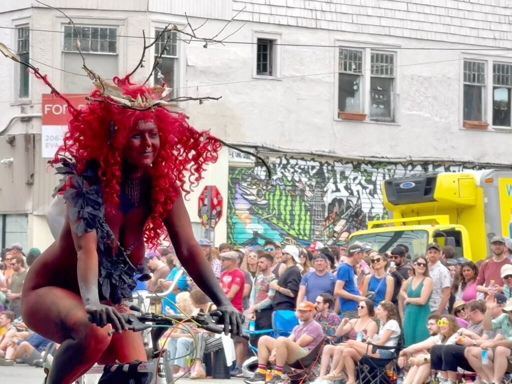 A vibrant performer in a red feathered costume rides a bicycle during a lively parade, with a crowd of spectators lined along the street.
