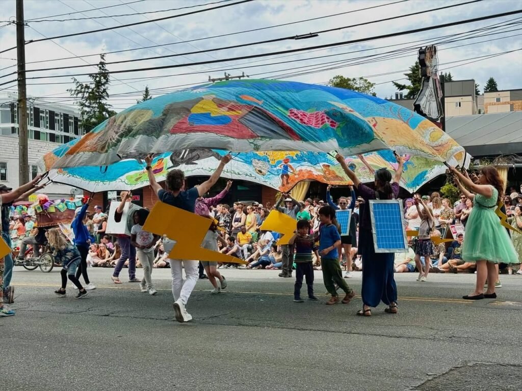 A group of people carries a large, colorful, and artistic canopy through a parade, engaging with a cheerful crowd.