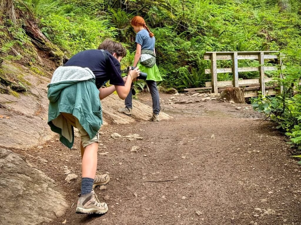 Two hikers on a dirt trail in a lush green forest with a wooden fence.
