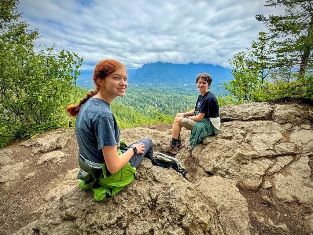Two people relaxing on a rocky hilltop with a panoramic mountain view in the Pacific Northwest.
