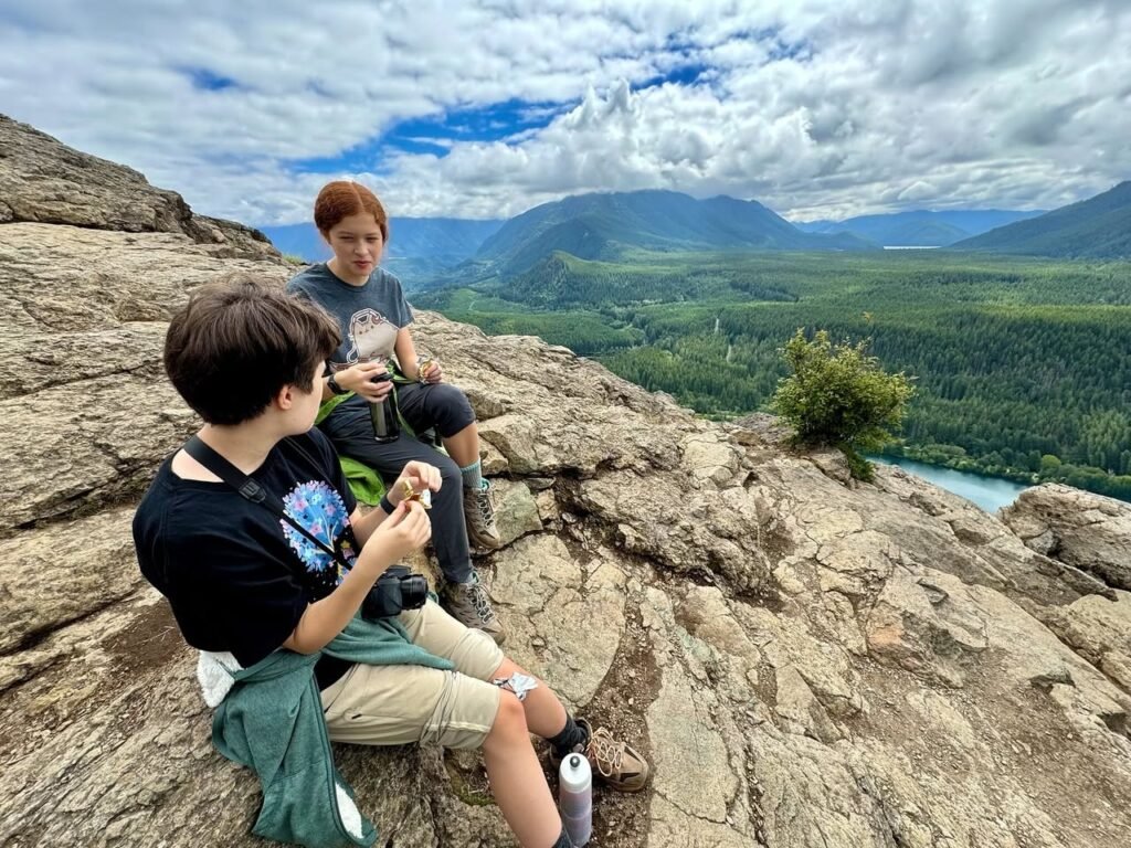 Hikers rest on rocky terrain overlooking forested mountains and a blue lake. A panoramic view of the Pacific Northwest landscape with cloud cover.