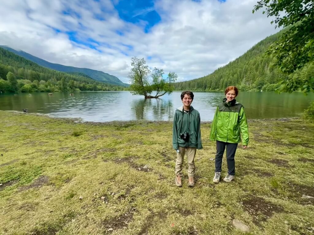 Two hikers standing on a grassy bank by a calm river in the Pacific Northwest, with forested mountains in the background.