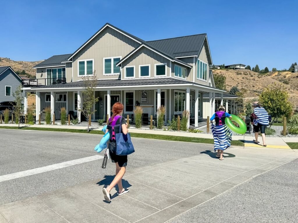 People carrying bags and swim gear walking past a modern lodge with a clear blue sky.