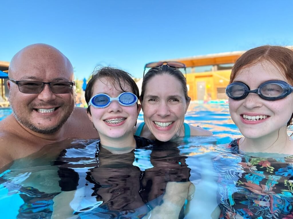 A family enjoys a sunny day at a water park pool, wearing swim goggles and smiling for the camera.