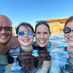 A family enjoys a sunny day at a water park pool, wearing swim goggles and smiling for the camera.