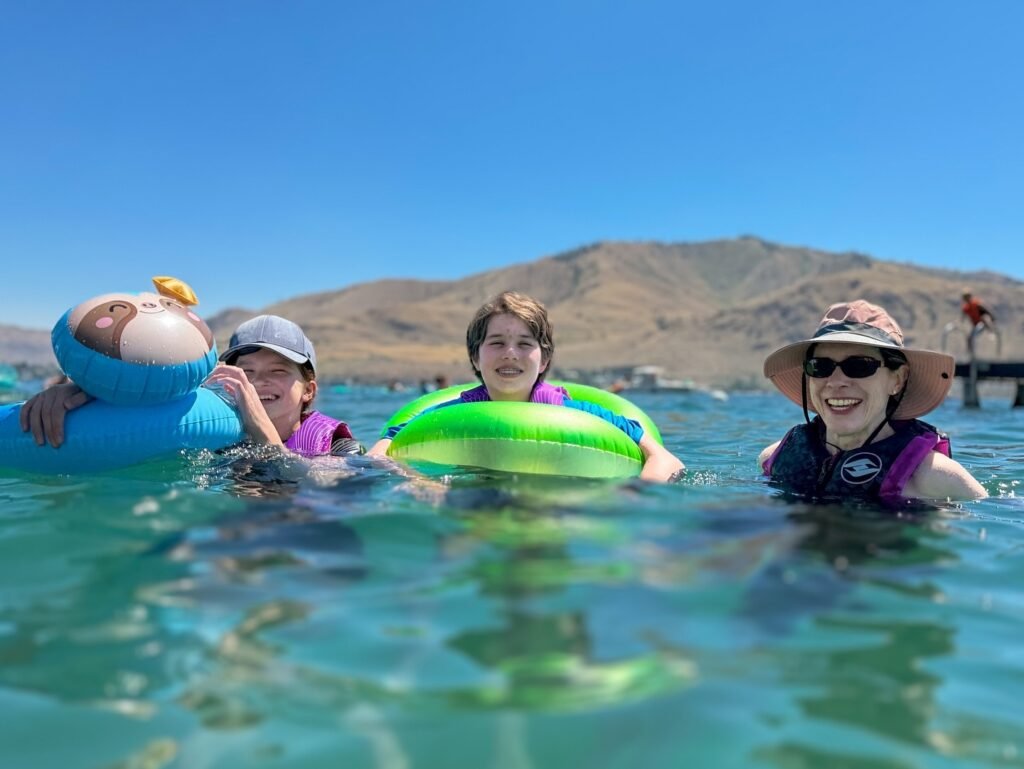 People enjoys summer in the water with colorful inflatable rings. The background features a clear blue sky and distant hills.