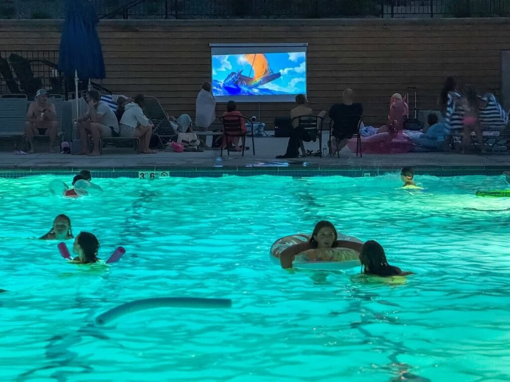 A swimming pool with people relaxing and a large outdoor screen showing a movie. The scene captures a casual evening of entertainment by the water.
