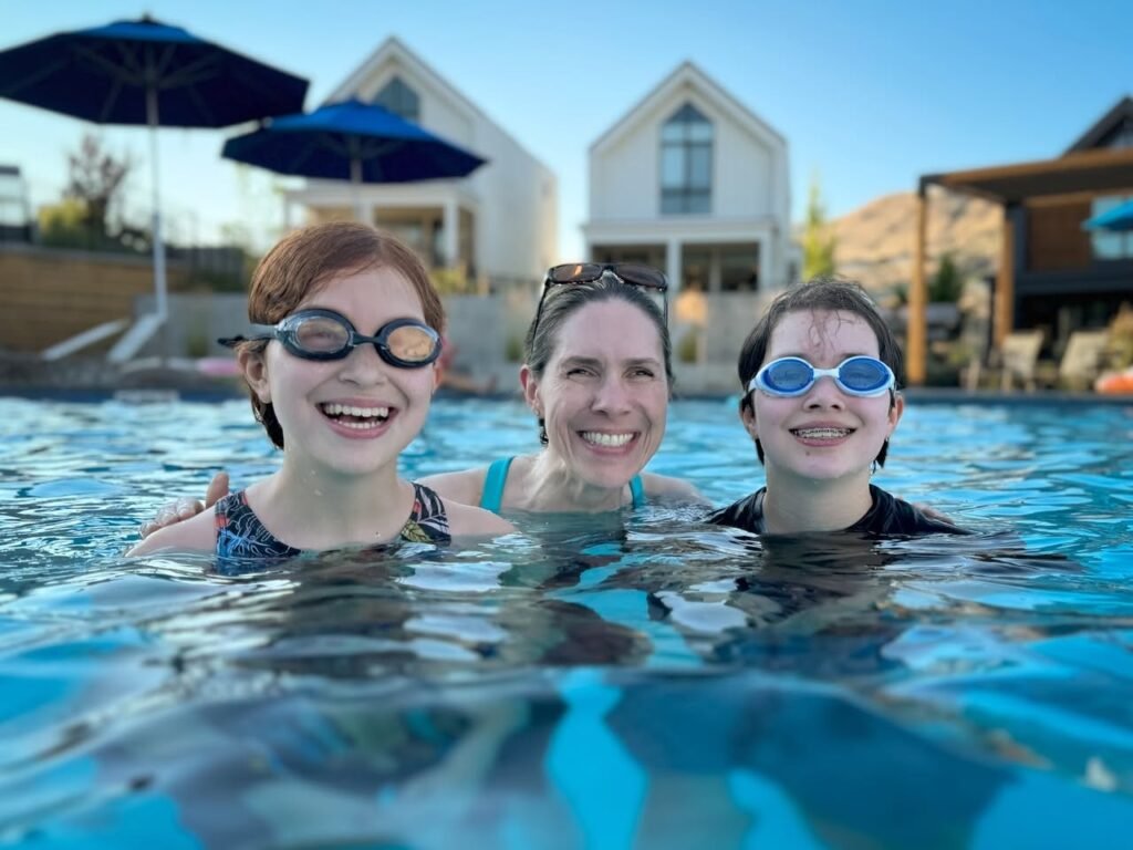 Three people wearing swimming glasses smiling in a clear blue outdoor pool with umbrellas and a stucco house in the background.
