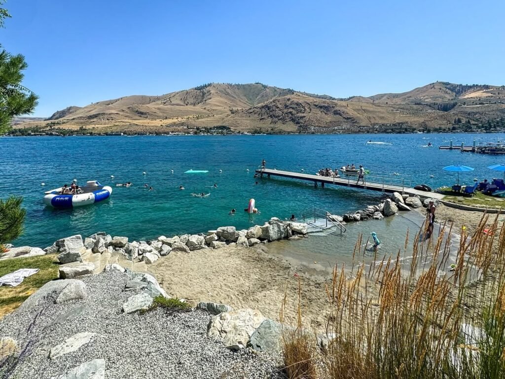 A sunny day at a lake with a sandy beach, clear blue water, and a mountain in the background.