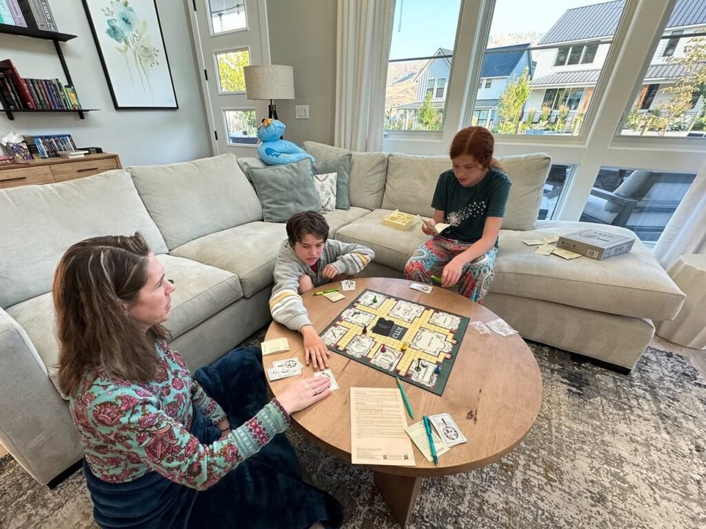 Family playing a board game at a round wooden table in a cozy living room. Natural light fills the space from large windows.