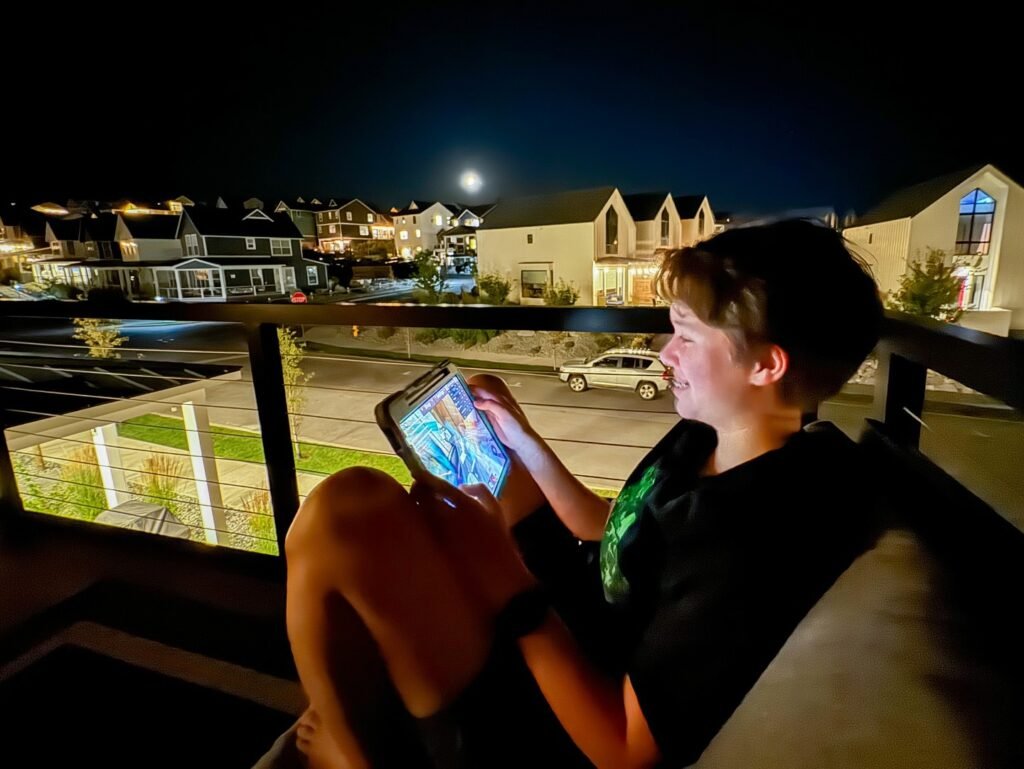 A person enjoys a tablet on a balcony at night. The view includes a moonlit sky and distant houses.