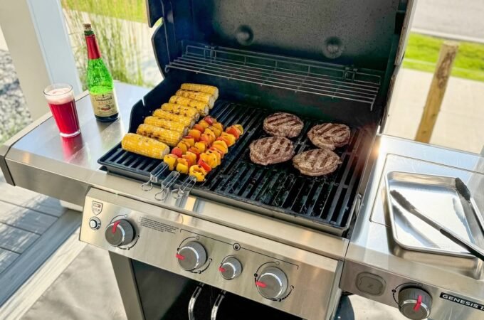 A stainless steel grill with burgers, corn, and vegetables cooking on the grates. Condiments and drinks are placed on the side.