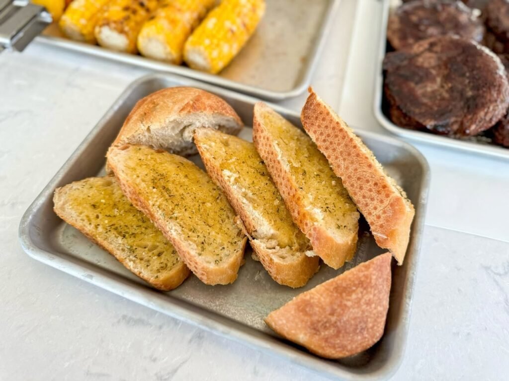 Slices of toasted garlic bread with herbs served on a metal tray alongside grilled corn on the cob. The rustic presentation suggests a shared outdoor dining experience in the Pacific Northwest.