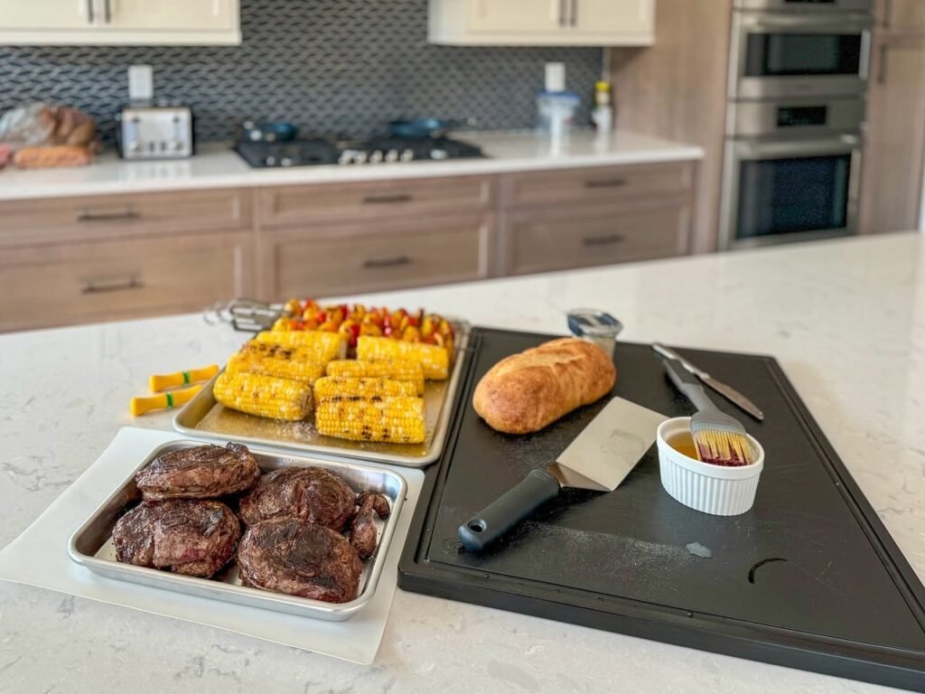 A spread of grilled corn, steaks, and bread on a cutting board in a modern kitchen. The food is arranged on a clean, white kitchen island counter.
