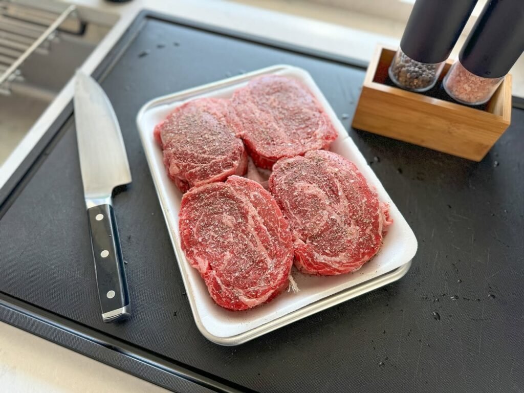 A close-up of raw steak patty in a white tray on a dark counter. A knife and seasoning caddie sit beside it.