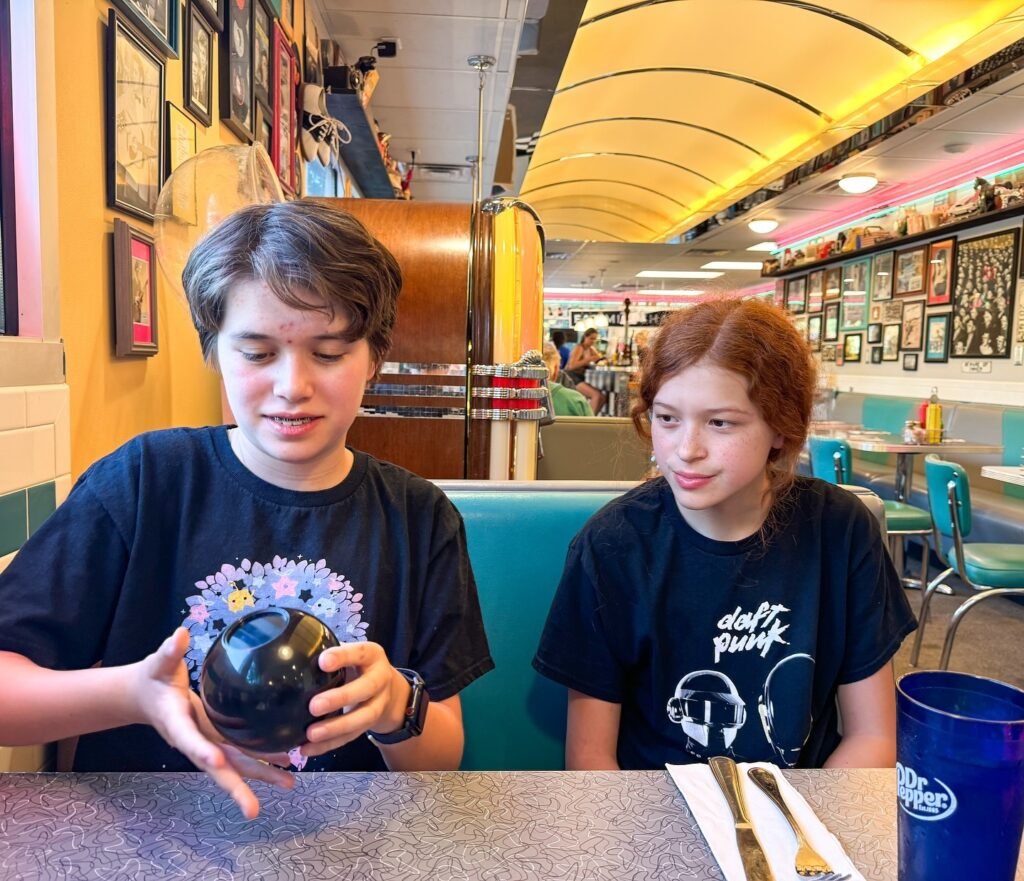 A young person plays with a black toy while seated at an In-N-Out burger restaurant table with another child. The setting includes teal booths and wooden paneling typical of the fast-food chain.