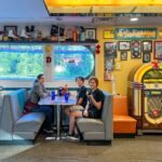 A family enjoys a meal at a retro 1950s diner in St. Paul, Minnesota. The interior features a classic jukebox, a surfboard, and vintage memorabilia on the shelves.