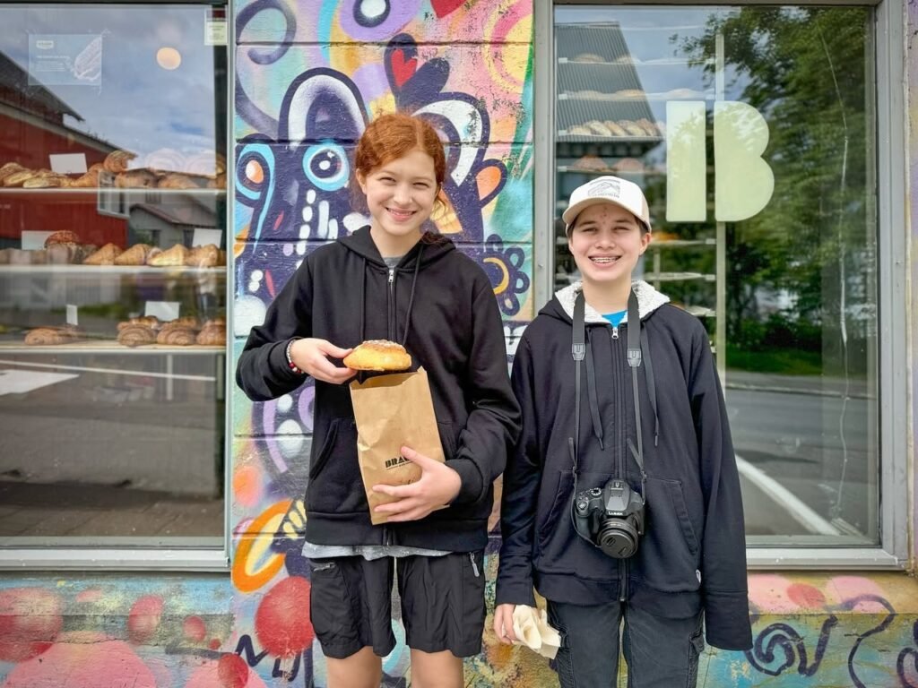 Two young people standing outside a bakery with colorful graffiti art, holding freshly baked goods in paper bags.