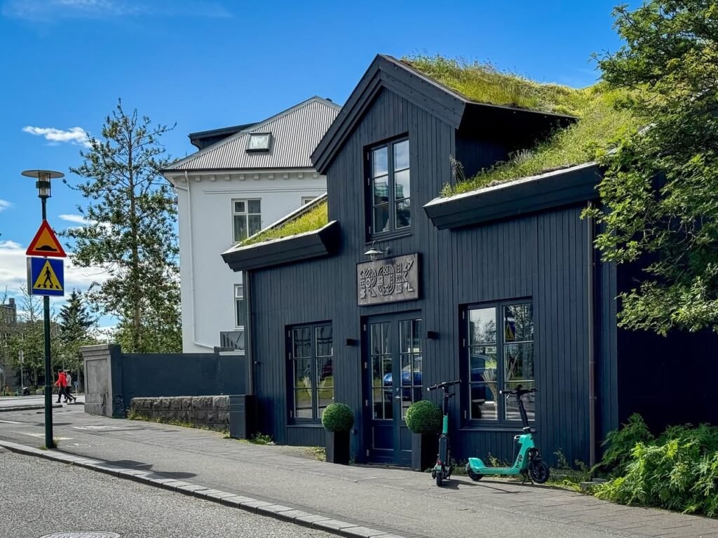 A charming Scandinavian street scene features a dark building with a lush green living roof and an electric scooter outside.