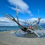 Þingvellir Visitors standing inside a large metal bird sculpture on a coastal promenade. The structure is set against a backdrop of clear blue sky and ocean.