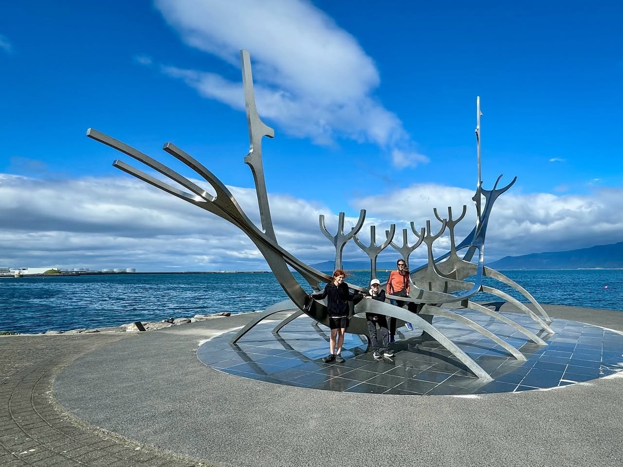 Reykjavík, Iceland Visitors standing inside a large metal bird sculpture on a coastal promenade. The structure is set against a backdrop of clear blue sky and ocean.